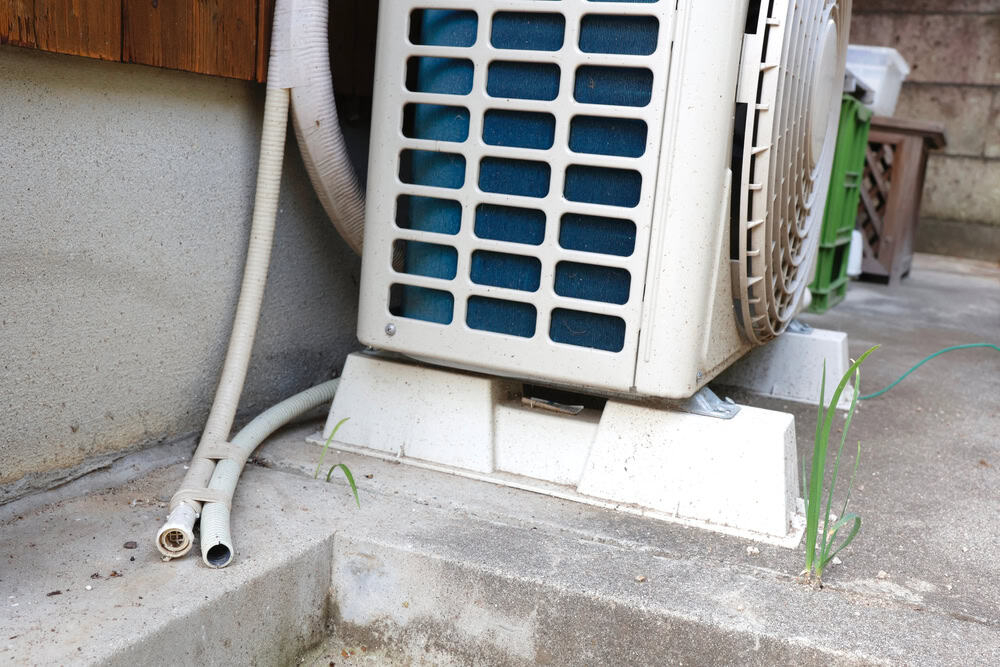 An outdoor air conditioning unit surrounded by concrete with a small plant sprouting nearby.