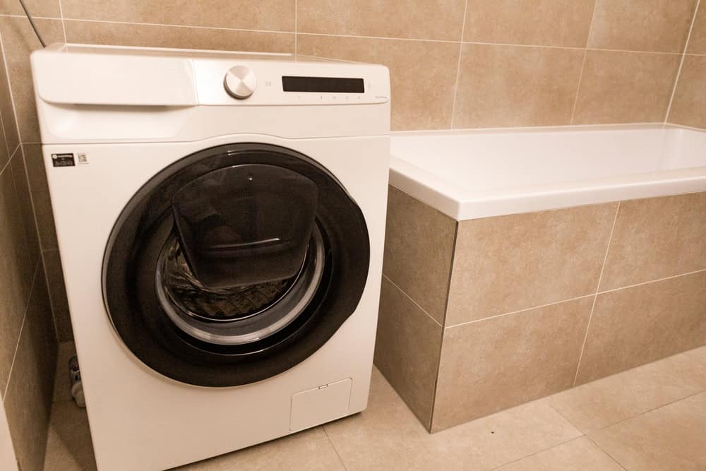 A modern washing machine stands next to a sleek bathtub in a stylish, tiled laundry-bathroom combo.