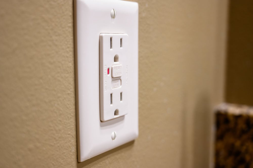 A close-up of a modern electrical outlet with a reset button, set against a beige wall.