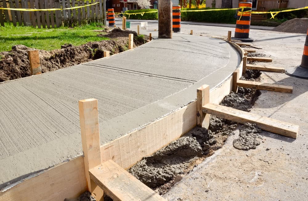 Freshly poured concrete sidewalk under construction, surrounded by wooden forms and traffic barriers.