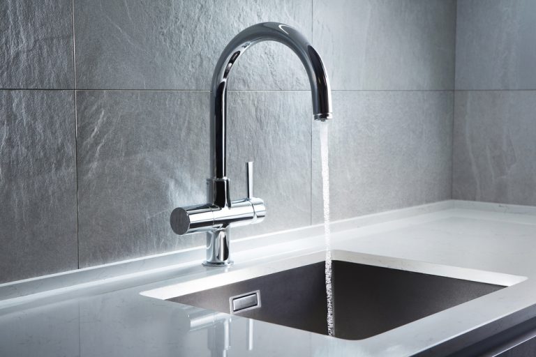 A sleek, modern kitchen faucet pours crystal-clear water into a black sink, set against a backdrop of elegant gray stone tiles.