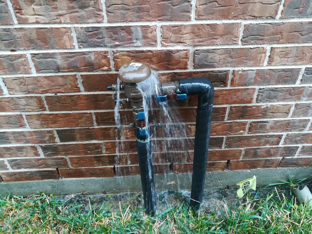 A broken outdoor water pipe sprays water against a brick wall, creating a small flood on the ground.