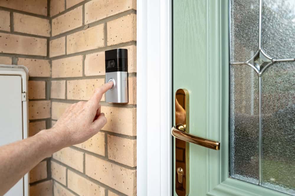 A hand presses a modern doorbell next to a green front door with a brass handle.