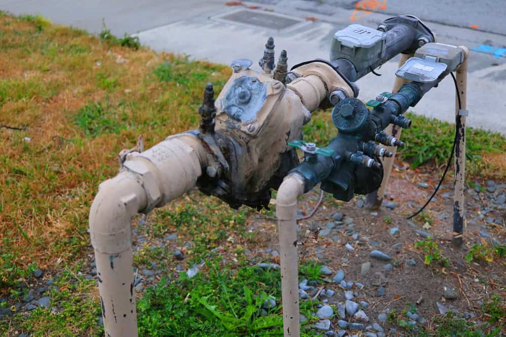 Rusty water meter and pipes on a patch of grass next to a sidewalk.