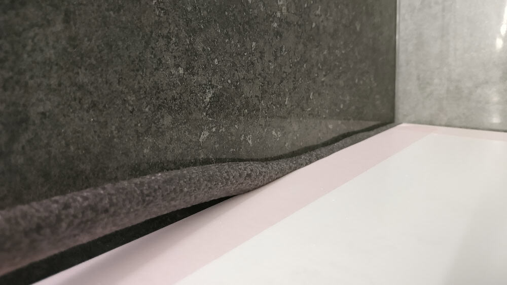 Bathroom sealant against grey textured tile and a white bathtub edge.