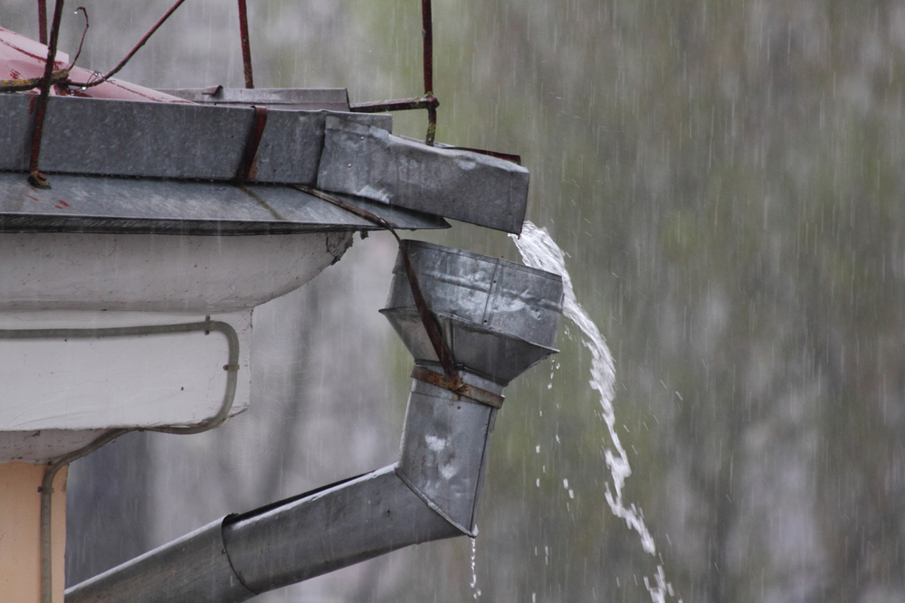 Rainwater cascades from a metal downspout during a heavy downpour, creating a graceful arc of water against the misty background.