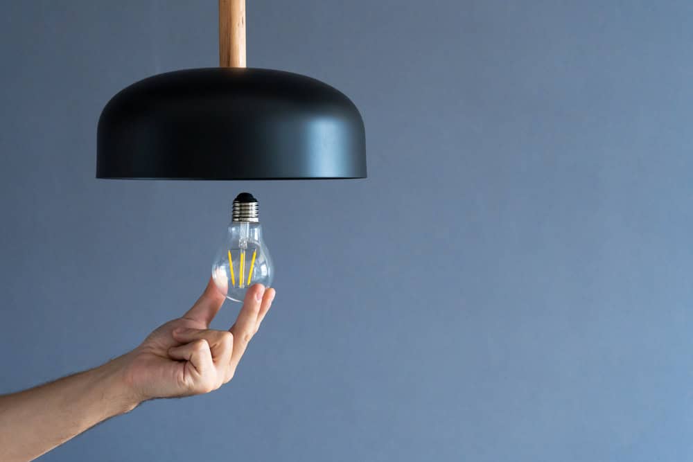 A hand delicately installs an LED bulb into a sleek black pendant lamp against a minimalist gray background.