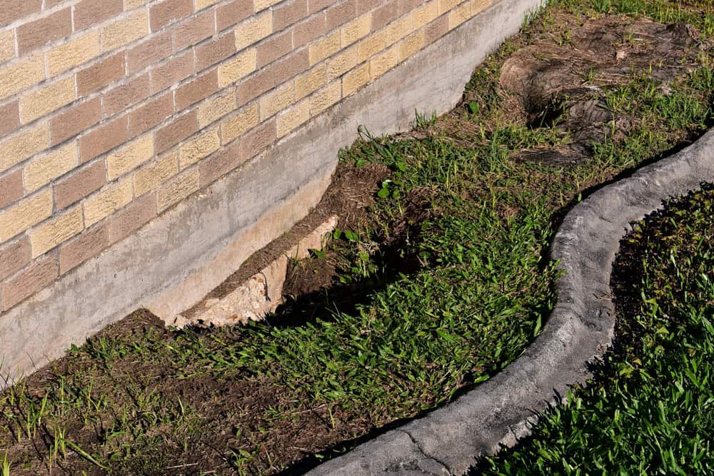 A cracked foundation exposes the vulnerability of a brick wall, surrounded by lush green grass.