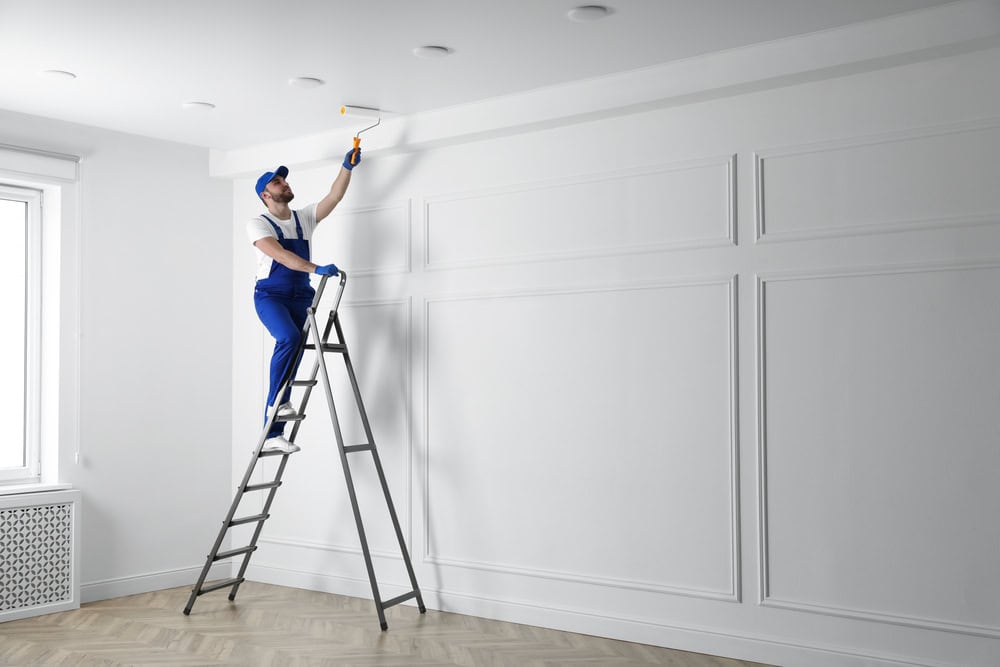 A painter meticulously works on the ceiling, standing on a ladder in a pristine white room.