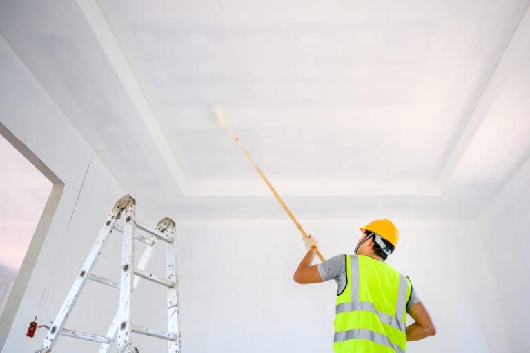 A construction worker in a high-visibility vest and hard hat paints the ceiling, standing on a ladder.
