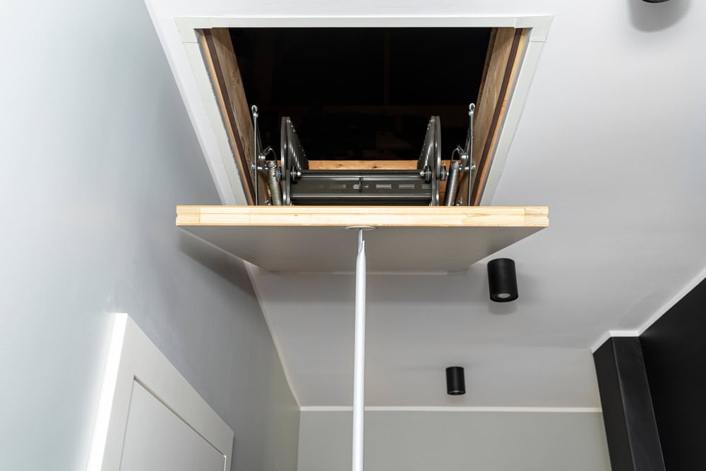 Retractable attic ladder partially extended from a ceiling hatch in a modern hallway.