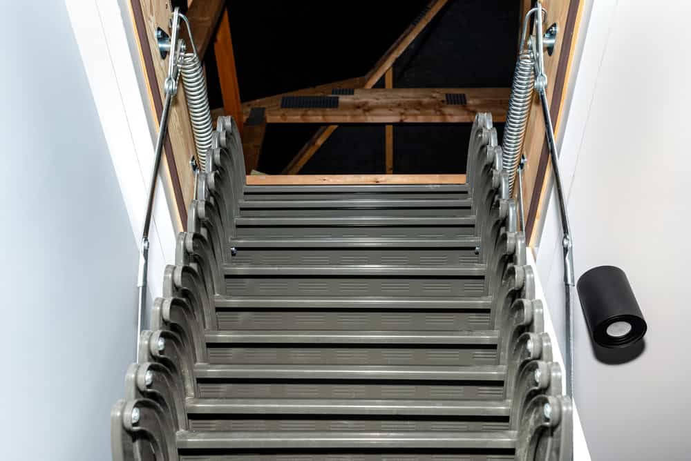 Retractable attic ladder leading up to a dark, unfinished loft space with exposed wooden beams.