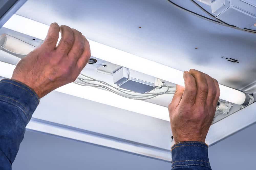 Installing LED lighting: A close-up of hands in denim sleeves mounting a fluorescent light fixture to a ceiling, with visible wiring and mounting brackets.