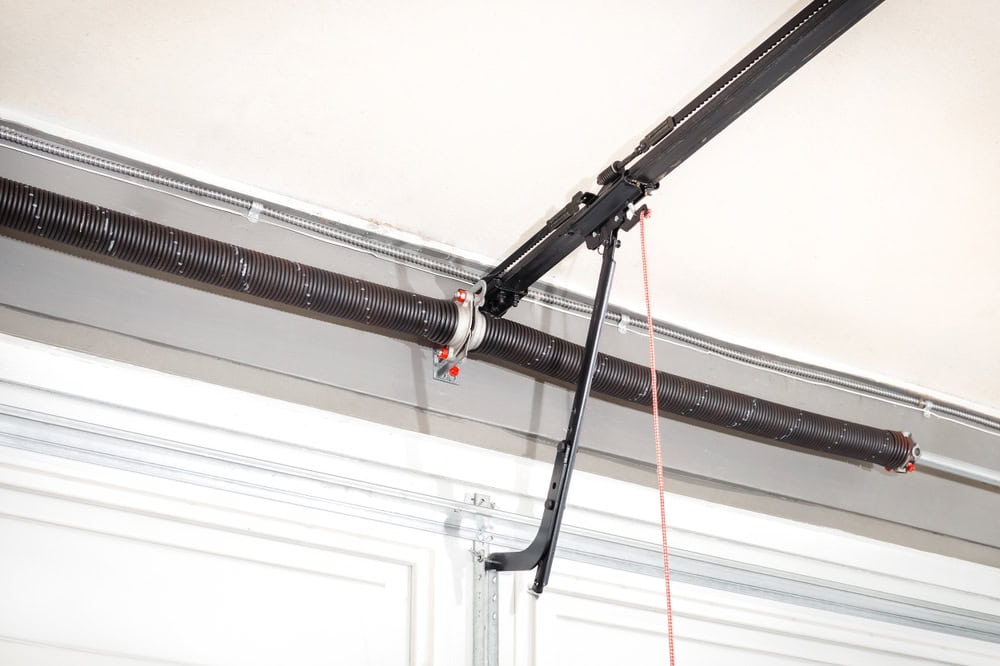 Garage door torsion spring and opener mechanism mounted above a sectional door, showing the black mounting bracket and safety cable installation.