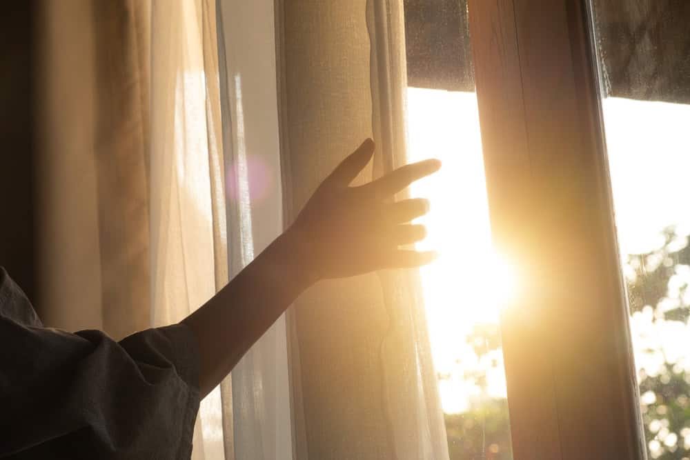 A hand reaches towards the sunlight streaming through a window, embracing the warmth of a new day.