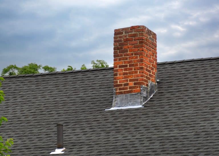 A classic red brick chimney rises from a shingled rooftop beneath a cloudy sky, framed by leafy green treetops.