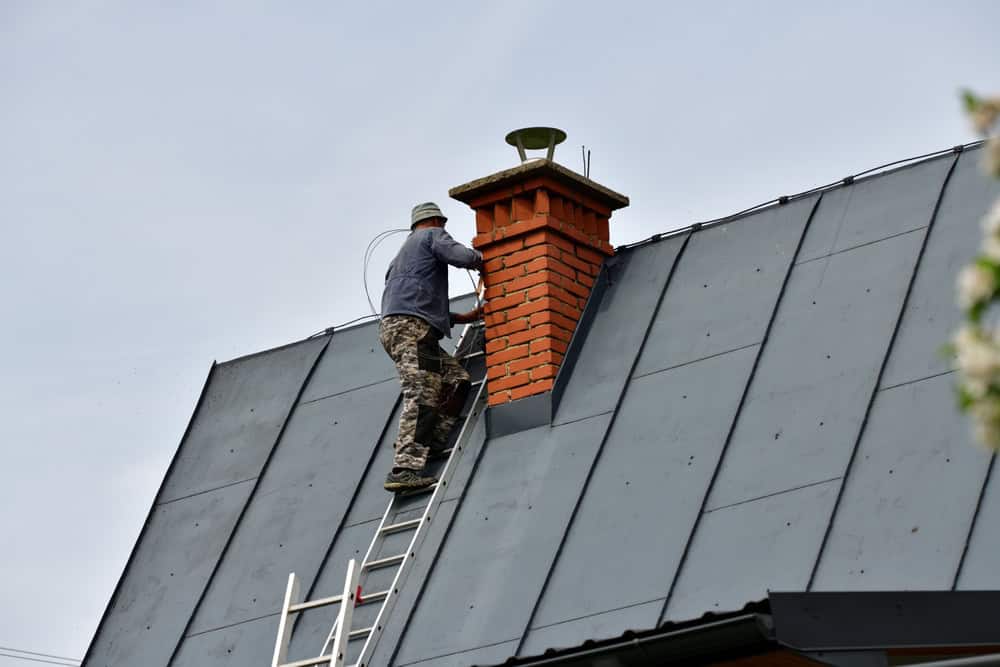Worker climbs onto a steep metal roof to inspect a red brick chimney under overcast skies.