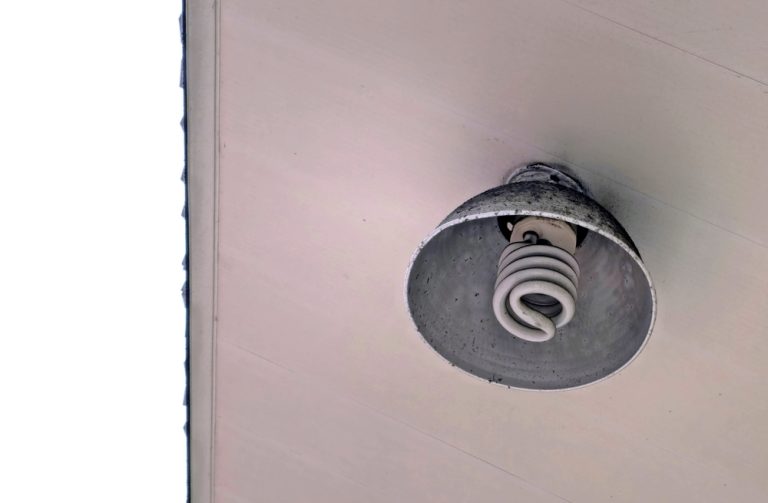 A weathered ceiling light fixture with a spiral CFL bulb, mounted beneath a covered outdoor roof.