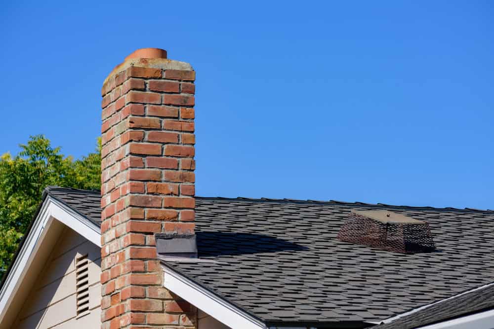 Brick chimney rising against clear blue sky, with asphalt shingles and gabled roofline of a residential home below