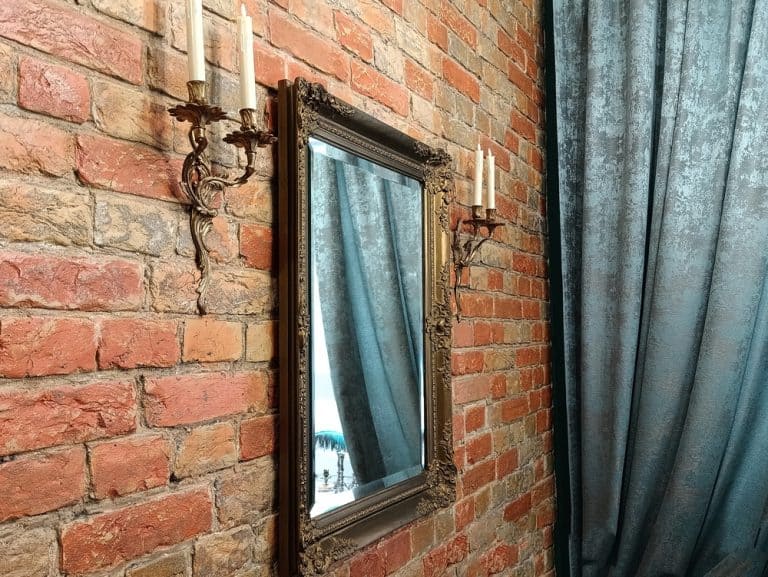 A vintage mirror reflects elegant blue curtains against a rustic brick wall adorned with ornate candle sconces.