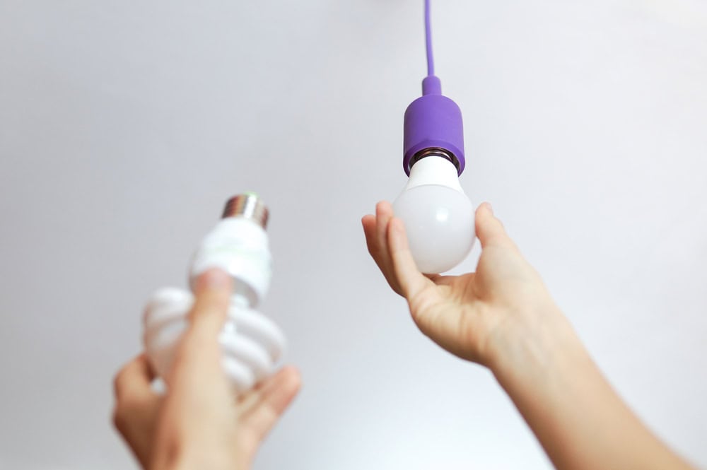 Hands replacing a CFL bulb with an LED under a modern purple socket.