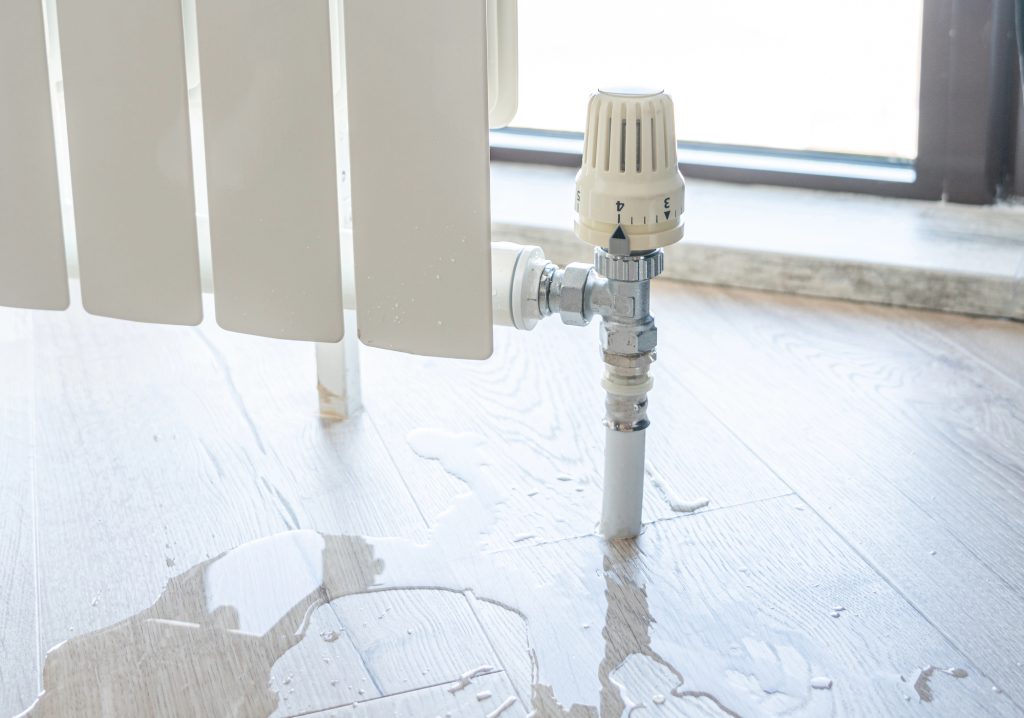 Leaking radiator valve creates a puddle on white wooden floor, highlighting the need for urgent maintenance