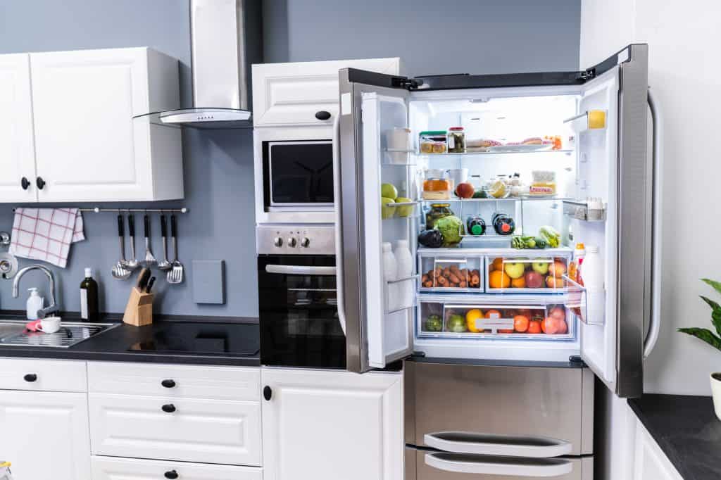 Modern white kitchen featuring an open stainless steel refrigerator stocked with fresh produce, alongside built-in appliances and sleek black countertops.