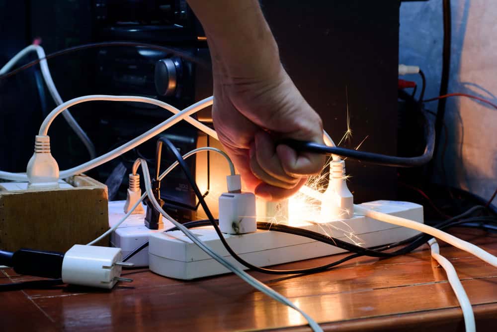A hand grabs a sparking power strip amidst a tangled mess of cables and devices.