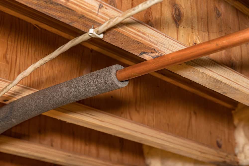 Exposed copper pipe with insulation in a rustic wooden attic.