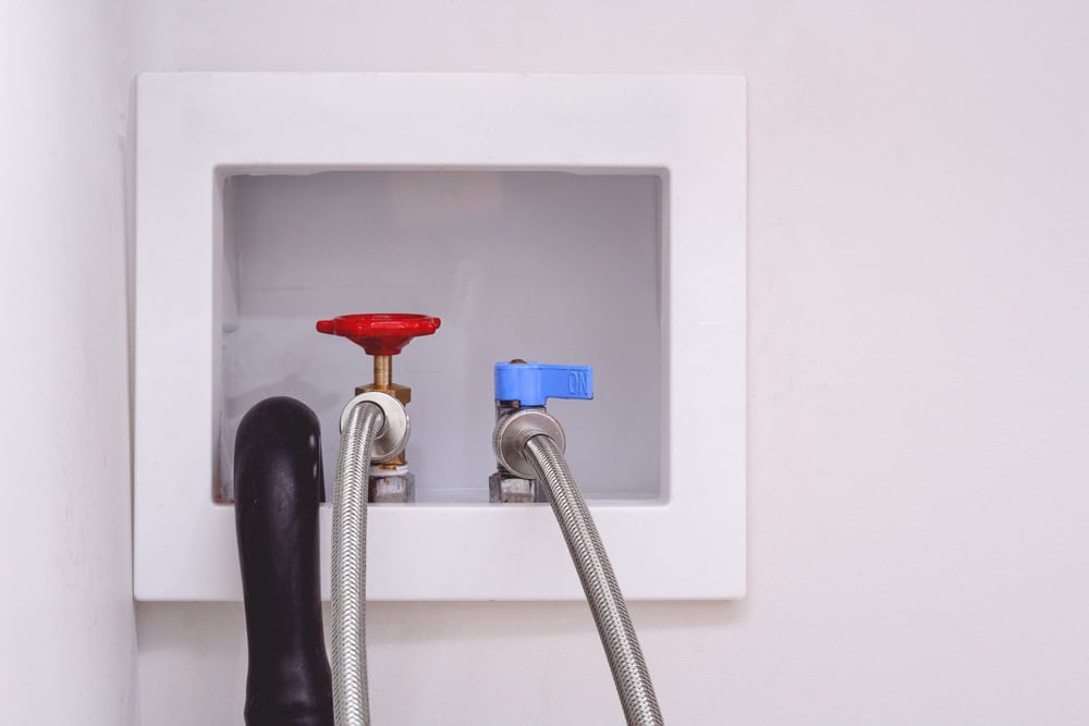 Washer hookup with hot (red) and cold (blue) water valves and a drain hose in a laundry room wall box.