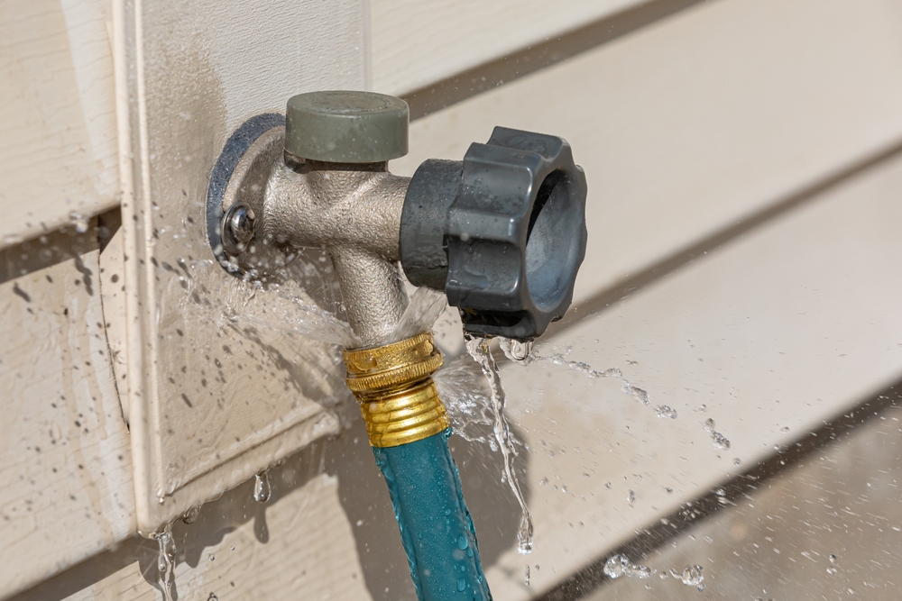 Leaky outdoor water spigot spraying water around a connected garden hose, signaling the need for repair or maintenance.