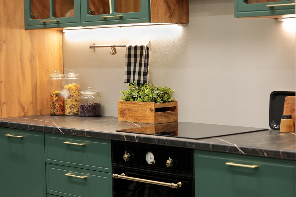 Modern kitchen featuring dark green cabinets, marble countertops, and gold hardware, accented with glass storage jars and a rustic wooden planter box with fresh herbs.