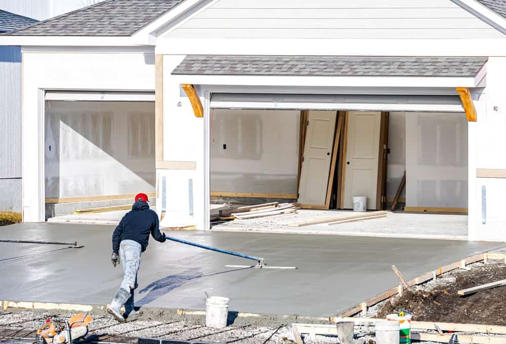Construction worker smoothing fresh concrete on a new residential driveway, with unfinished garage bays visible in the background