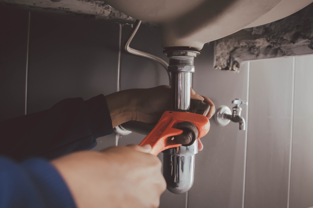 Plumber's hands grip an adjustable wrench while working on a chrome sink trap beneath a bathroom vanity.