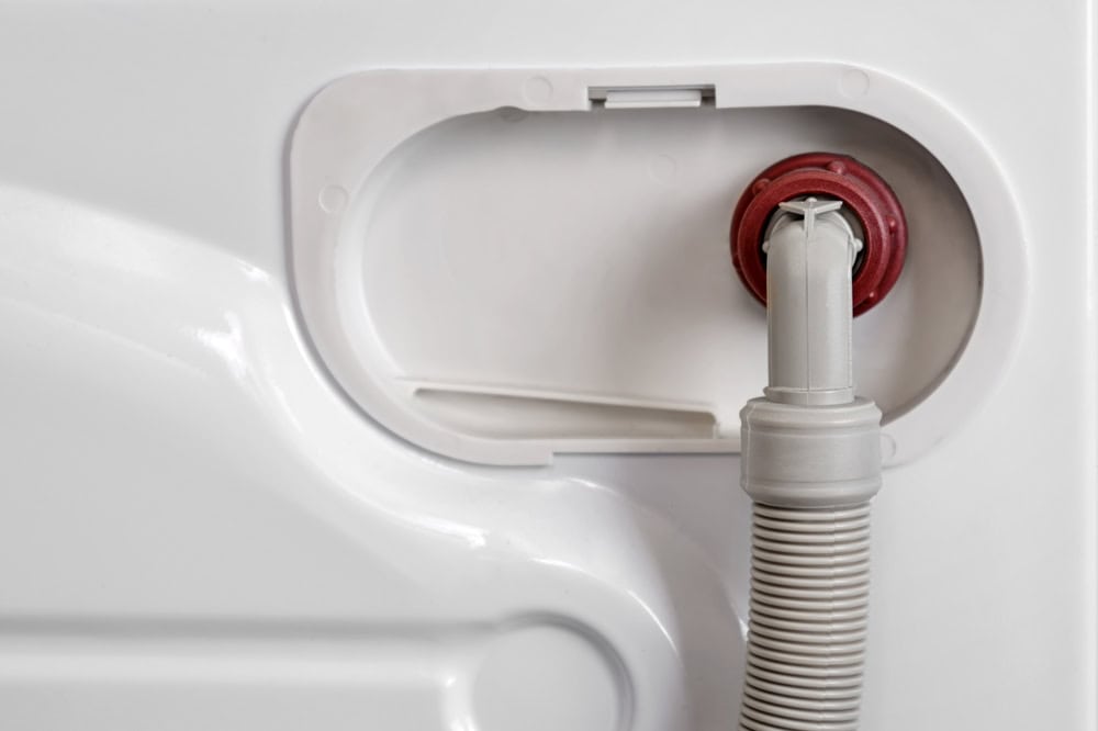 Inside a washing machine drain hose connection point, showing a white corrugated drain hose with a red seal mounted in a recessed access panel.