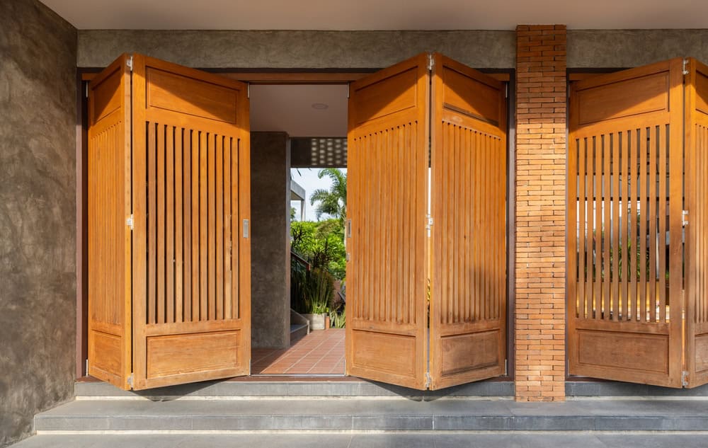 Warm wooden folding doors open to a sunlit tropical courtyard.