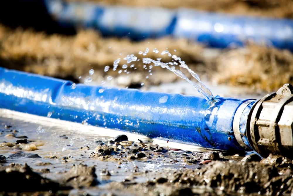 Water leaks from a damaged blue hose, highlighting the urgency of infrastructure repair in a muddy field.