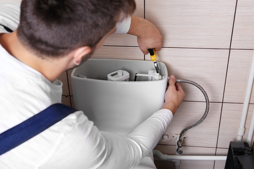 Plumber repairing toilet tank mechanism with screwdriver, wearing white long-sleeve shirt