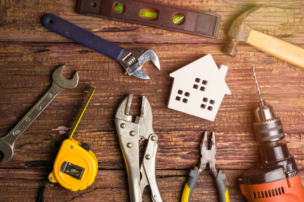 Home Repair Essentials: A collection of tools including wrenches, pliers, a level, hammer, measuring tape, and power drill arranged around a miniature white house model on a rustic wooden surface.