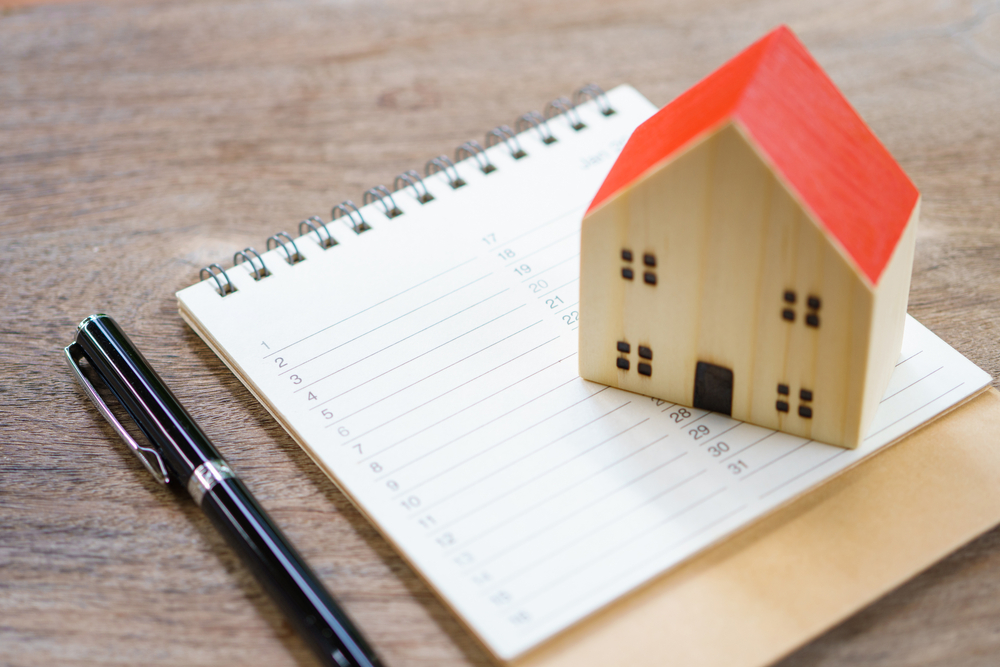 A miniature wooden house with a bright red roof sits atop a spiral-bound calendar or planner, with a sleek black pen placed alongside. This clean, minimalist composition suggests real estate planning, mortgage scheduling, or home-related appointments on a wooden desk surface.