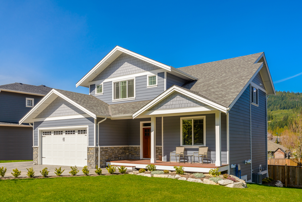 Modern craftsman-style home featuring gray siding, stone accents, and a welcoming front porch with seating, set against a blue mountain backdrop.