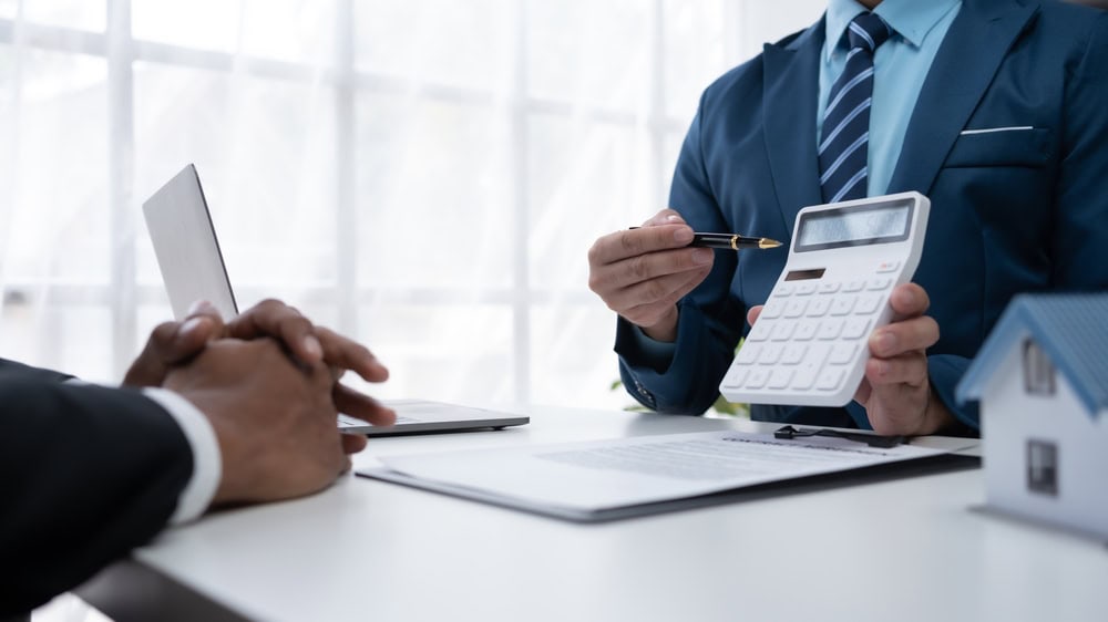 A real estate agent discusses figures with a client, laptop and model house on the table.