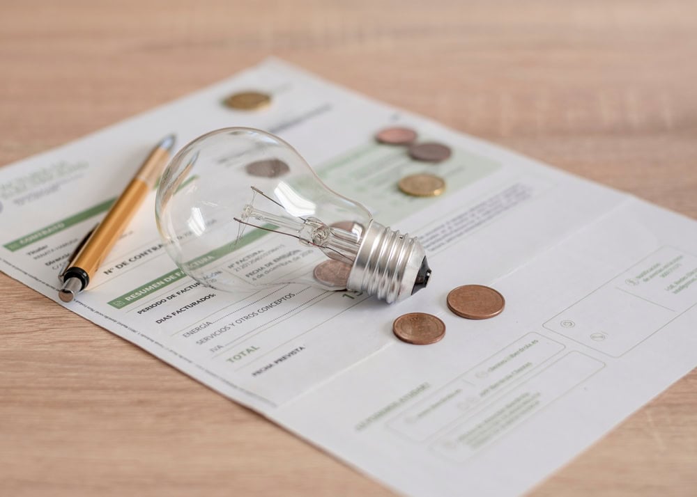 A light bulb resting on an electricity bill with coins scattered around.