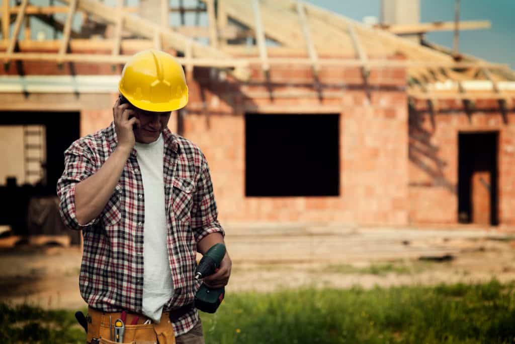Construction worker in plaid shirt and yellow hard hat takes a phone call while holding a power drill at a residential building site