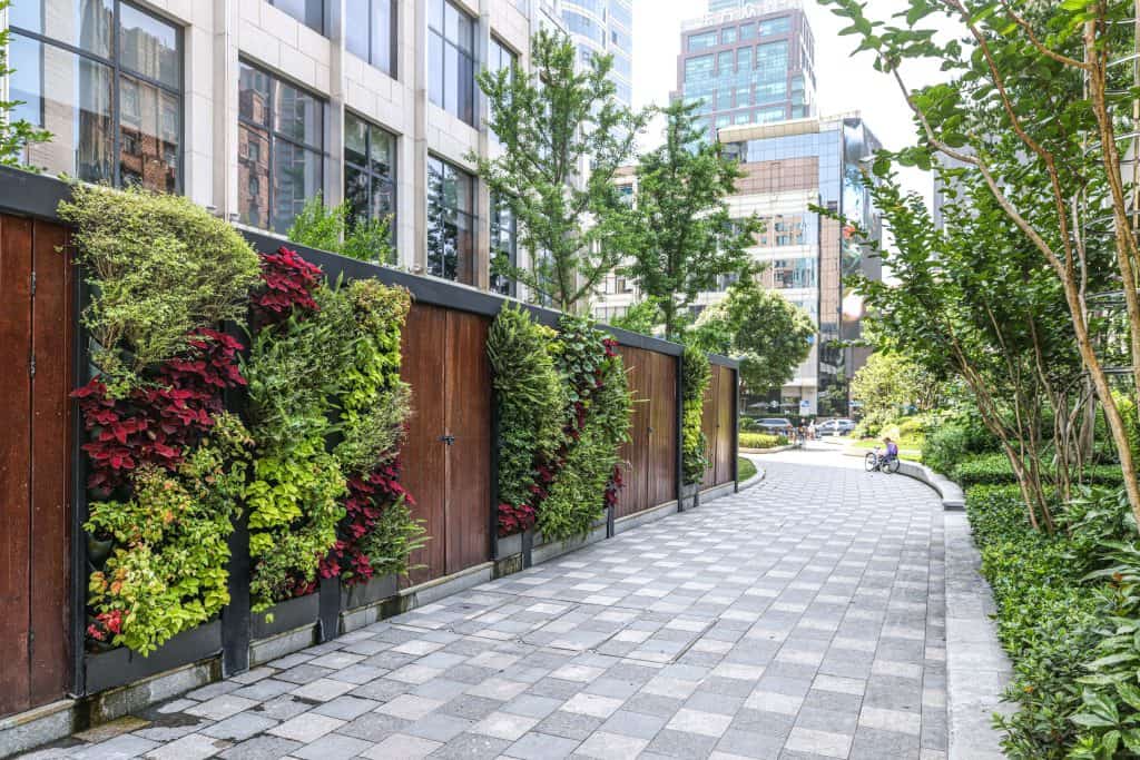A vibrant outdoor vertical garden integrated into a wooden fence along a modern city walkway. The garden features a variety of colorful plants, including red and green foliage, adding natural beauty to the urban setting. Surrounding trees and shrubs complement the landscaped area, while contemporary buildings rise in the background under a bright sky. A cyclist rests in the distance, enhancing the peaceful atmosphere.