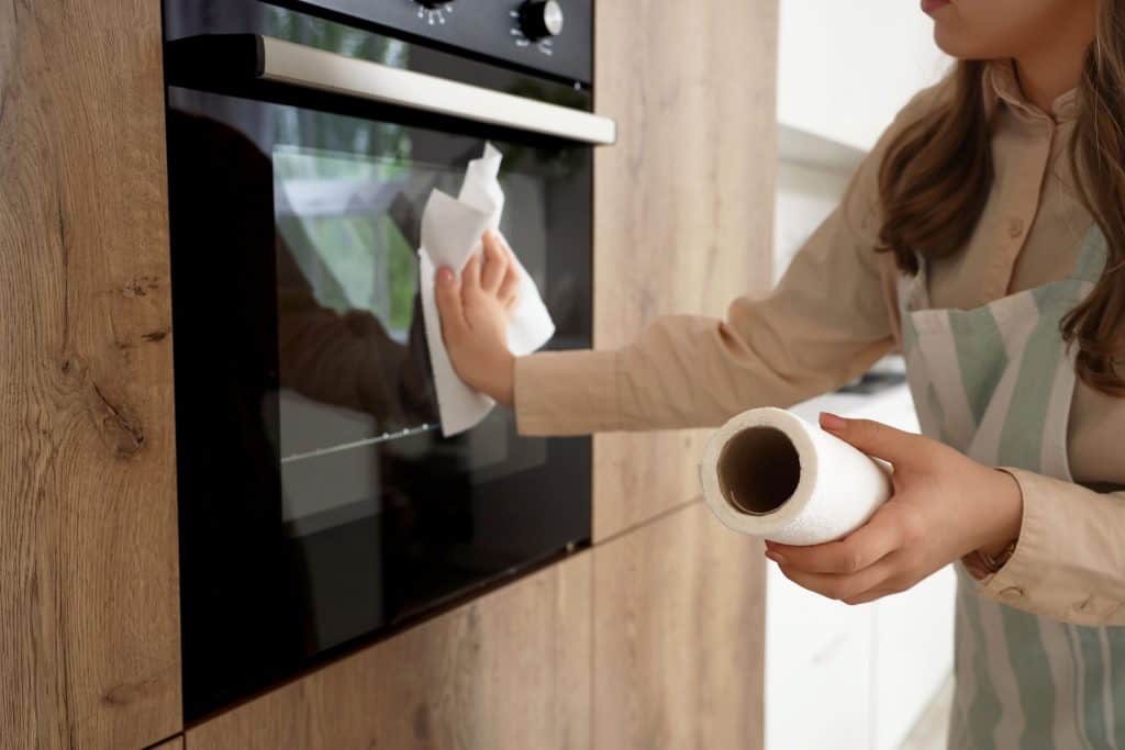 A woman wearing an apron cleans the glass door of a modern oven in a wooden-finish kitchen. She uses a paper towel in one hand while holding the roll in the other, ensuring a spotless surface.