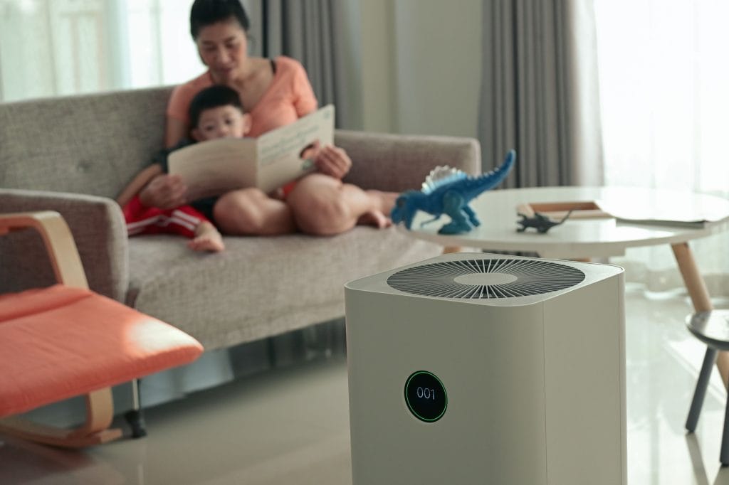 A modern air purifier with a digital display runs in the foreground of a cozy living room, while a mother reads a book to her child on the couch. A blue toy dinosaur sits on a white table near the window, creating a peaceful and family-oriented atmosphere. Caption: Keeping indoor air clean for a healthier family environment – a quality air purifier enhances comfort and reduces pollutants in your home.