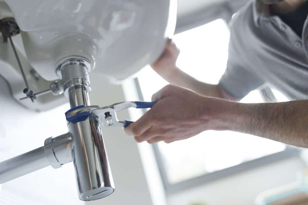 A plumber uses a wrench to repair the piping beneath a bathroom sink. The shiny chrome pipe contrasts with the soft lighting from a nearby window, highlighting the attention to detail during the fix.
