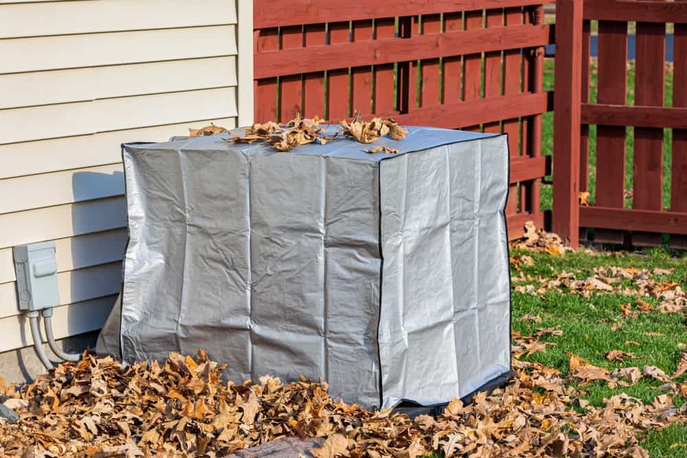 A backyard air conditioning unit wrapped for winter protection, surrounded by fallen autumn leaves.