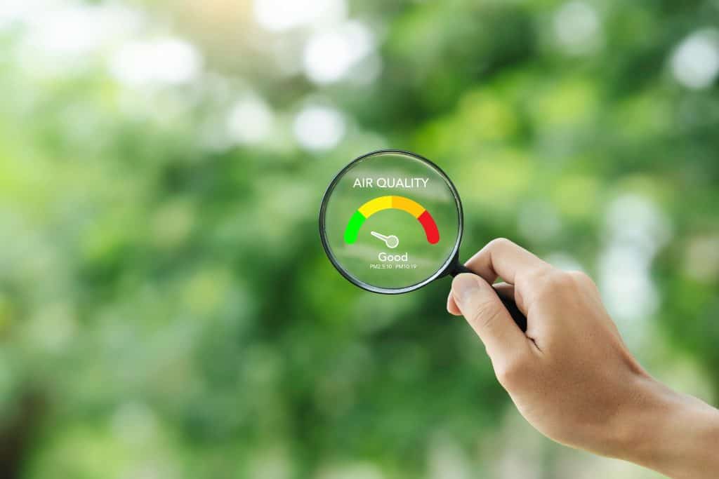 A hand holds a magnifying glass displaying an air quality meter showing "Good," set against a blurred background of green foliage. The meter uses a color scale from green to red, symbolizing air quality levels.